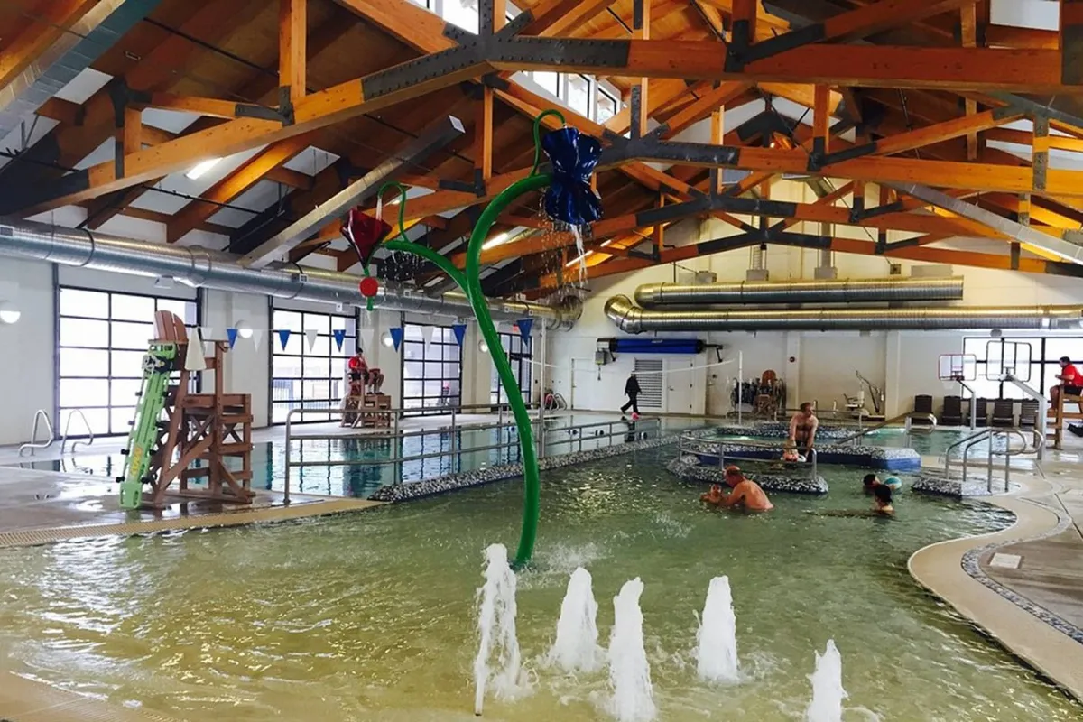 A large, vibrant indoor pool featuring colorful water features, slides, and a spacious play area under exposed wooden beams. The lively environment shows families and children enjoying aquatic activities.