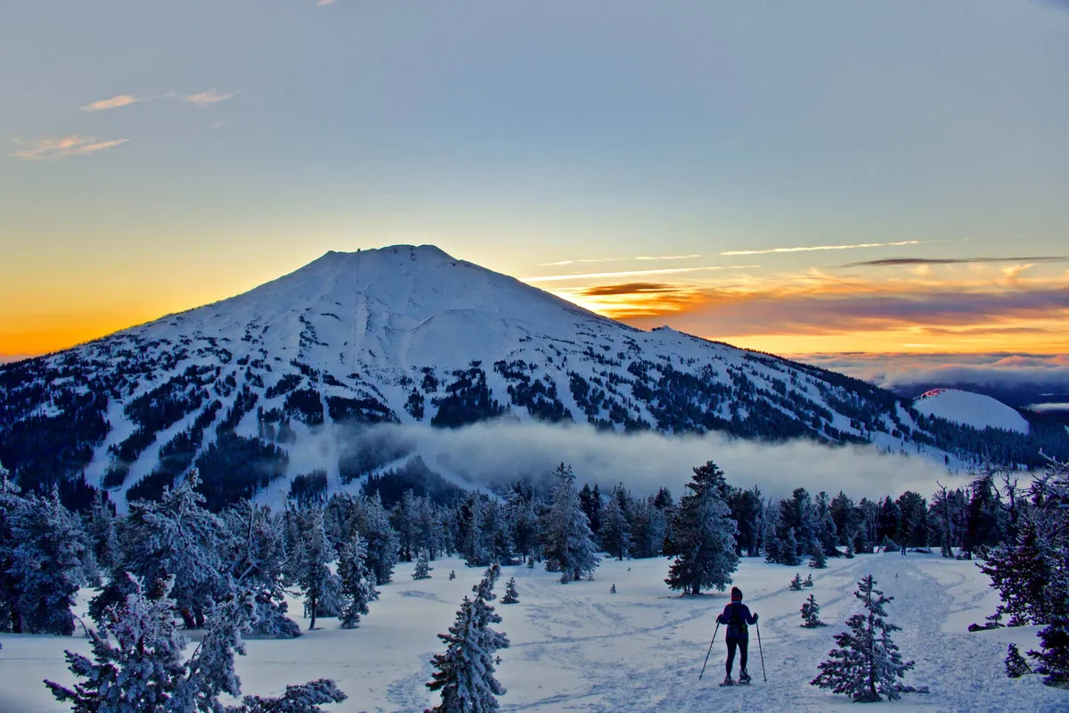 A dramatic mountain landscape at sunset, with a skier descending a snow-covered slope. The backdrop showcases snow-capped peaks and a glowing sky, setting a scene of winter sports excitement and scenic splendor.