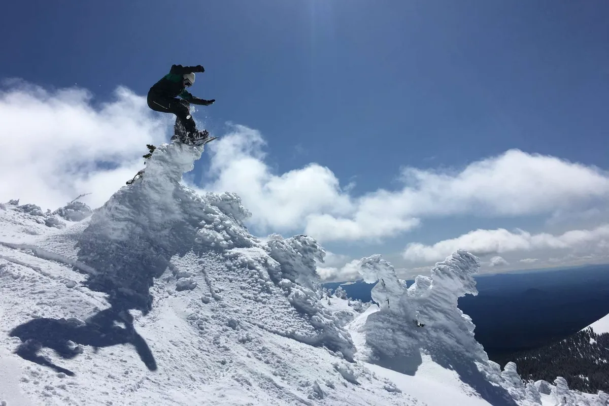 An exhilarating shot of a snowboarder carving down a snow-covered mountain peak, with expansive blue skies and clouds in the background. The scene captures the thrill and natural beauty of winter sports near Sunriver, with the majestic slopes of Mt. Bachelor visible.