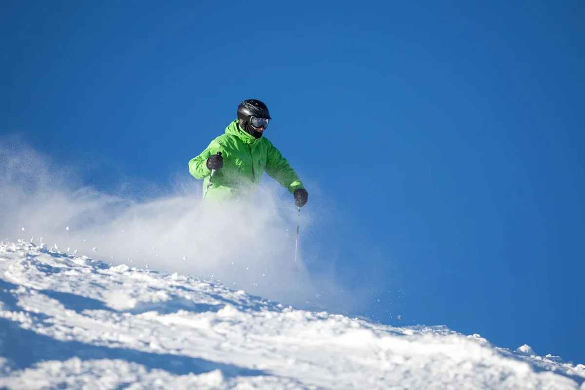 A skier gracefully descends a pristine, snow-covered slope against a bright blue sky. The composition emphasizes speed, excitement, and the magnificent mountain environment that surrounds Sunriver.