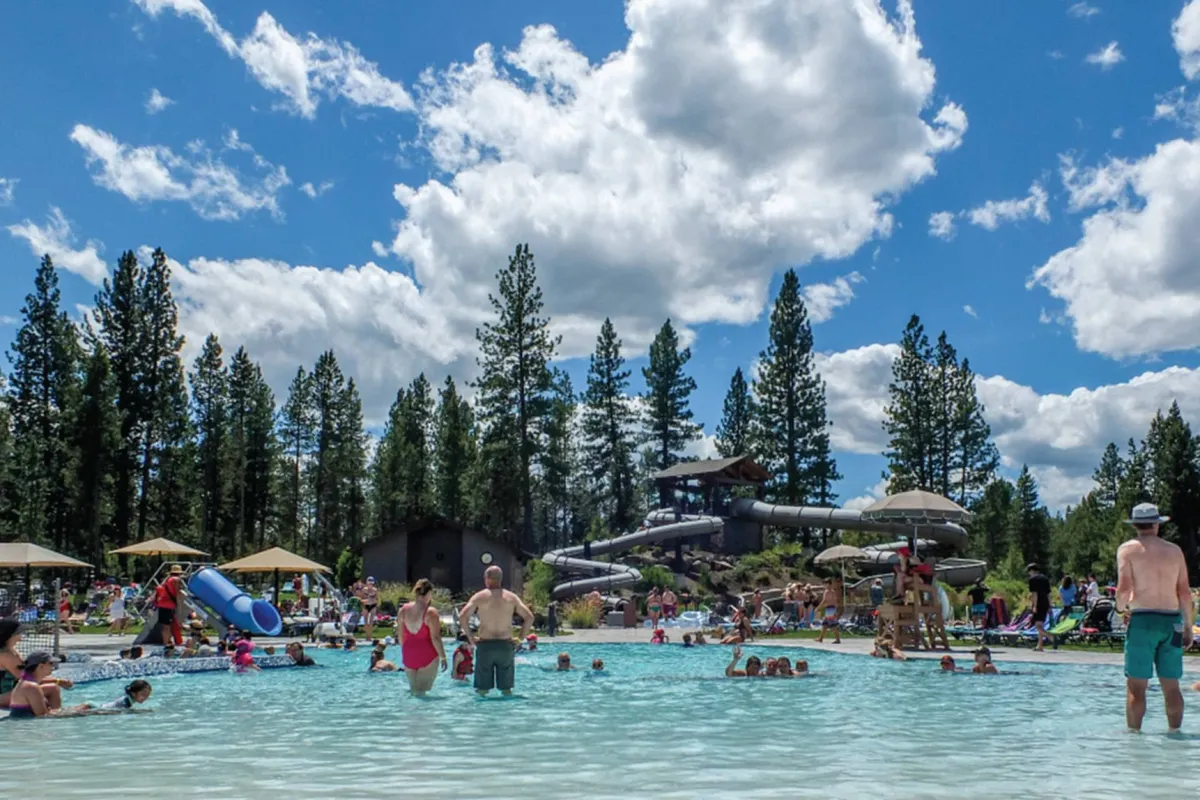 An outdoor swimming pool scene with a water slide, tall pine trees in the background, families relaxing under sun umbrellas, and people socializing and swimming. The mood is energetic and joyful.