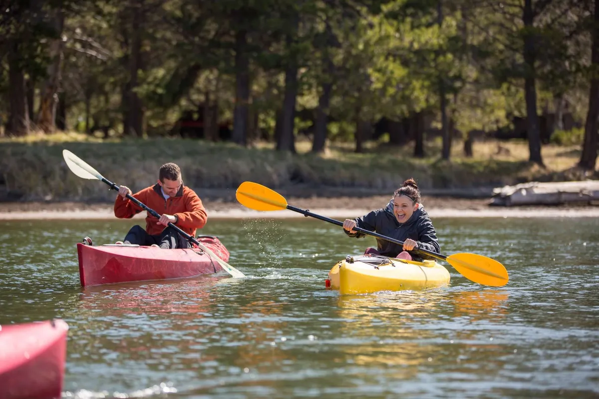 An upbeat scene of two colorful kayaks—red and yellow—floats on a sparkling river, with participants smiling and paddling. The water's surface reflects the vibrant kayaks, emphasizing the thrill of kayaking and bonding over outdoor sports.