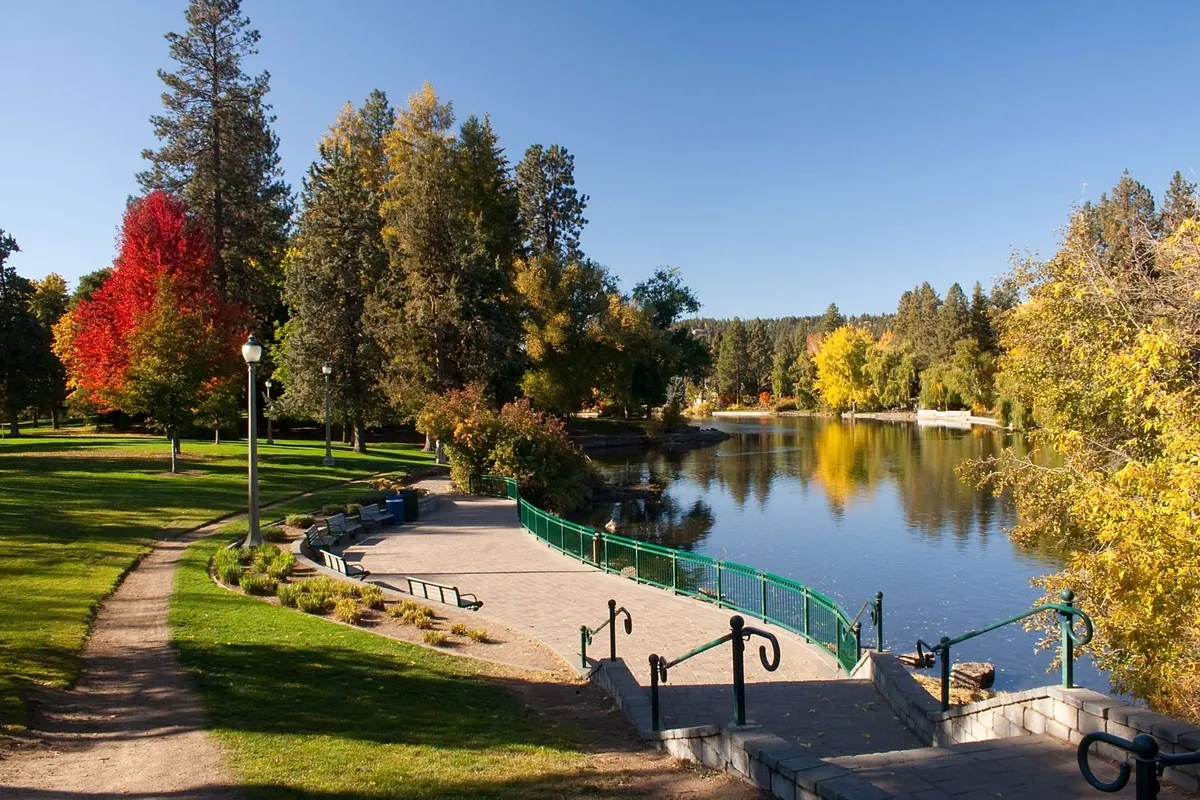 A tranquil exterior scene showcases a calm water surface bordered by colorful autumn trees. A wooden walkway, park benches, and an arched bridge invite visitors to walk, jog, picnic, or simply enjoy the beauty of the outdoors. The setting radiates peace and offers glimpses of local wildlife.