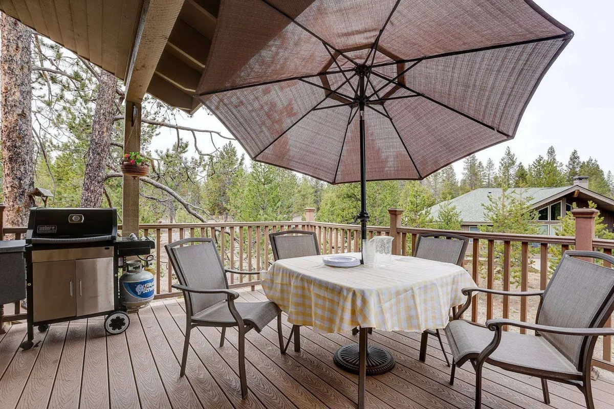 A spacious outdoor deck featuring a table with a checkered tablecloth, a large umbrella for shade, and a grill—perfect for al fresco dining. The setting is relaxed and inviting, suggesting a welcoming space for meals and gatherings.