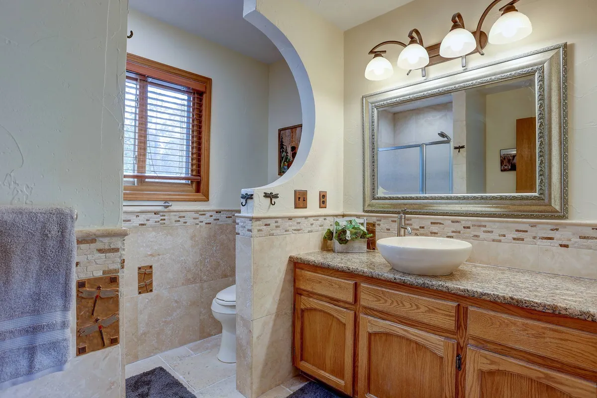 A serene bathroom featuring a granite countertop, distinctive sink, large mirror, and warm wooden cabinetry, with a window letting in natural light. The calm tones evoke a spa-like atmosphere.