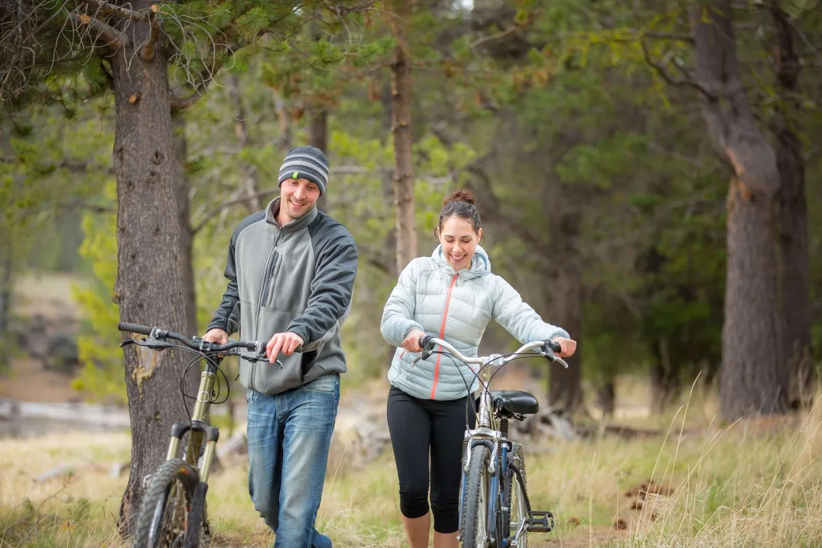 Two people walk together with bicycles through a lush, open area framed by tall trees and grassy fields. The relaxed outdoor setting evokes a sense of exploration and shared adventure in nature.