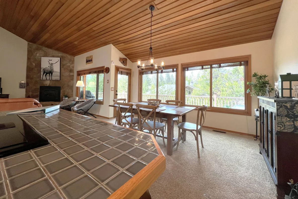 A spacious dining area with a sturdy wooden table and chairs sits next to large windows, offering a bright space for meals. The fireplace and exposed wooden beams add character and warmth, while a decorative lamp provides a homey touch. The room sets the stage for memorable family meals and celebrations.