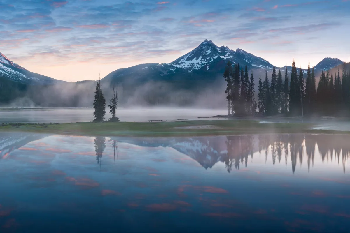 A breathtaking view of mountains, a serene lake, and tall trees under an open sky. The natural beauty invites outdoor exploration and relaxation, echoing the nearby Sunriver cafés, spa, and nature center highlighted in the narrative.
