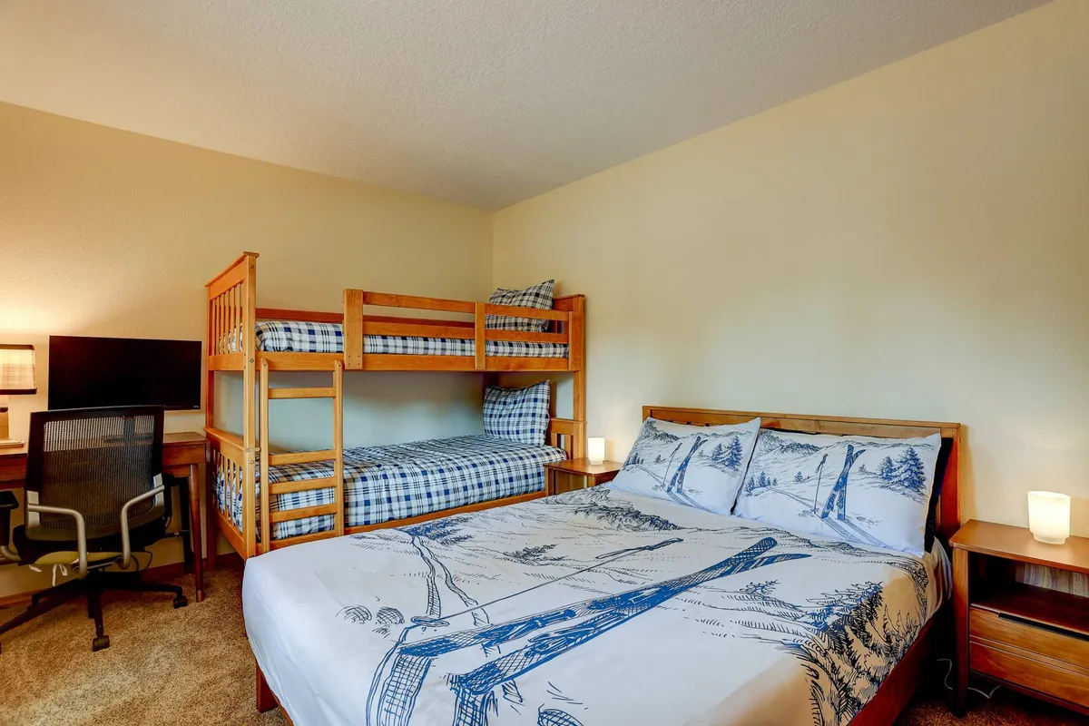 A family-friendly room featuring a bunk bed, double bed, study desk with a computer monitor, and a nightstand with a lamp. The presence of a workstation makes it suitable for remote work or online learning, as highlighted in the section.