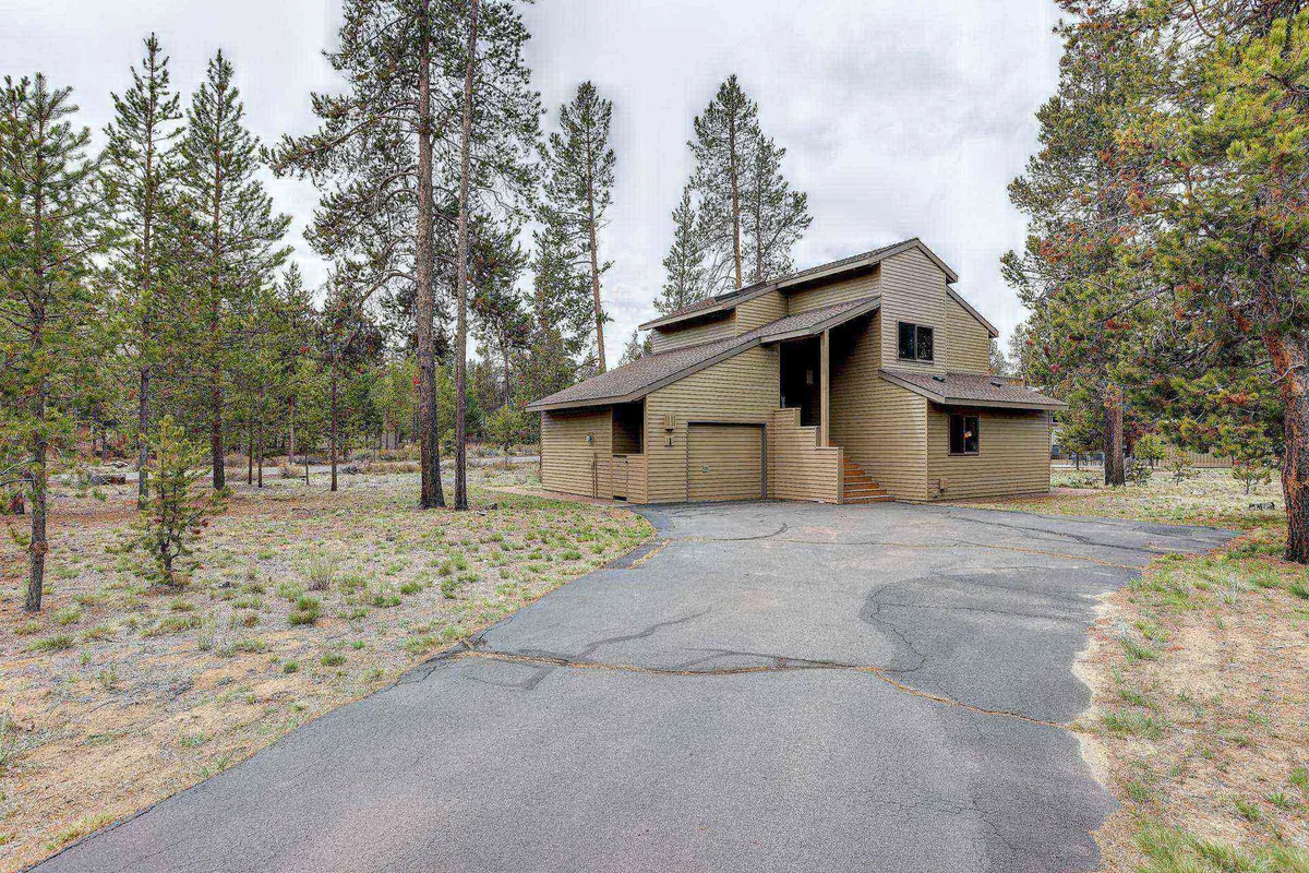 The image showcases the exterior of a charming single-family home nestled among tall trees, with a spacious driveway leading up to the house. The setting is serene, capturing the warm glow of sunset filtering through the forest, creating an inviting and tranquil atmosphere.