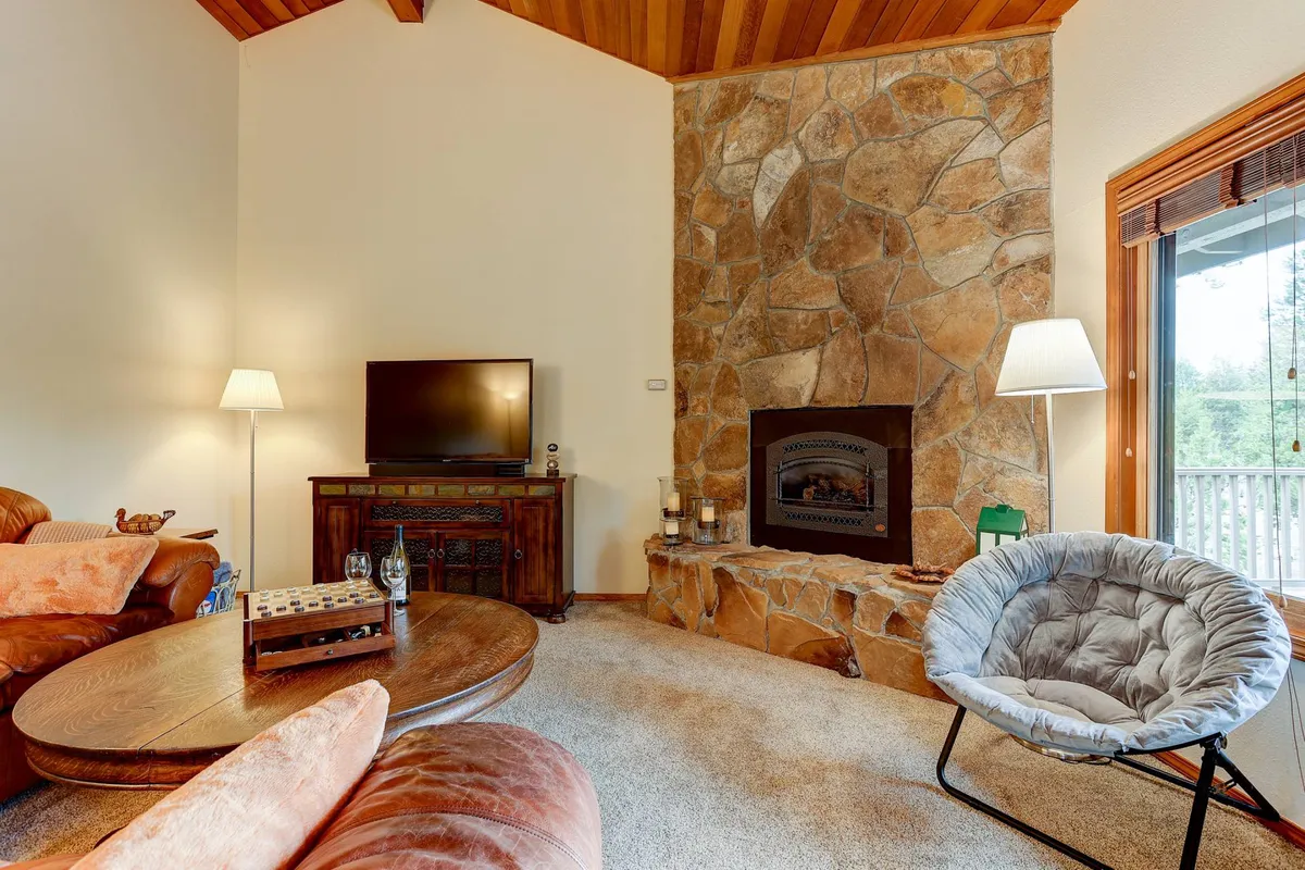 A cozy living room with a stone fireplace as the focal point, surrounded by comfortable seating and wooden furniture. Large windows allow natural light to pour in, creating a bright, welcoming atmosphere. The setup encourages relaxation, conversation, and family gatherings, all centered around the warmth of the fireplace.