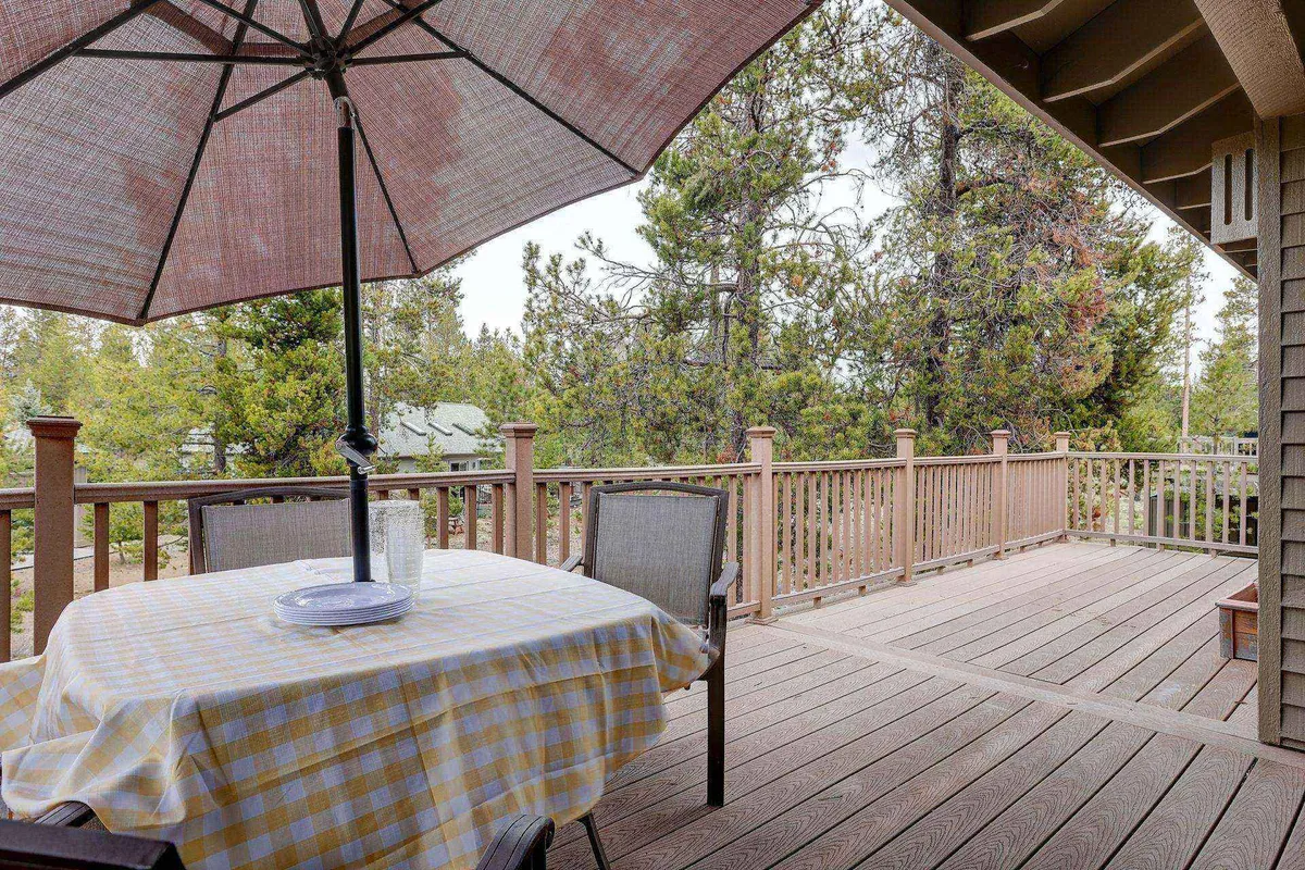 A spacious outdoor deck featuring a table with a checkered tablecloth under a large umbrella, with deck railing and tall pine trees visible in the background. The space is set up for outdoor dining, reading, and relaxing while immersed in nature.