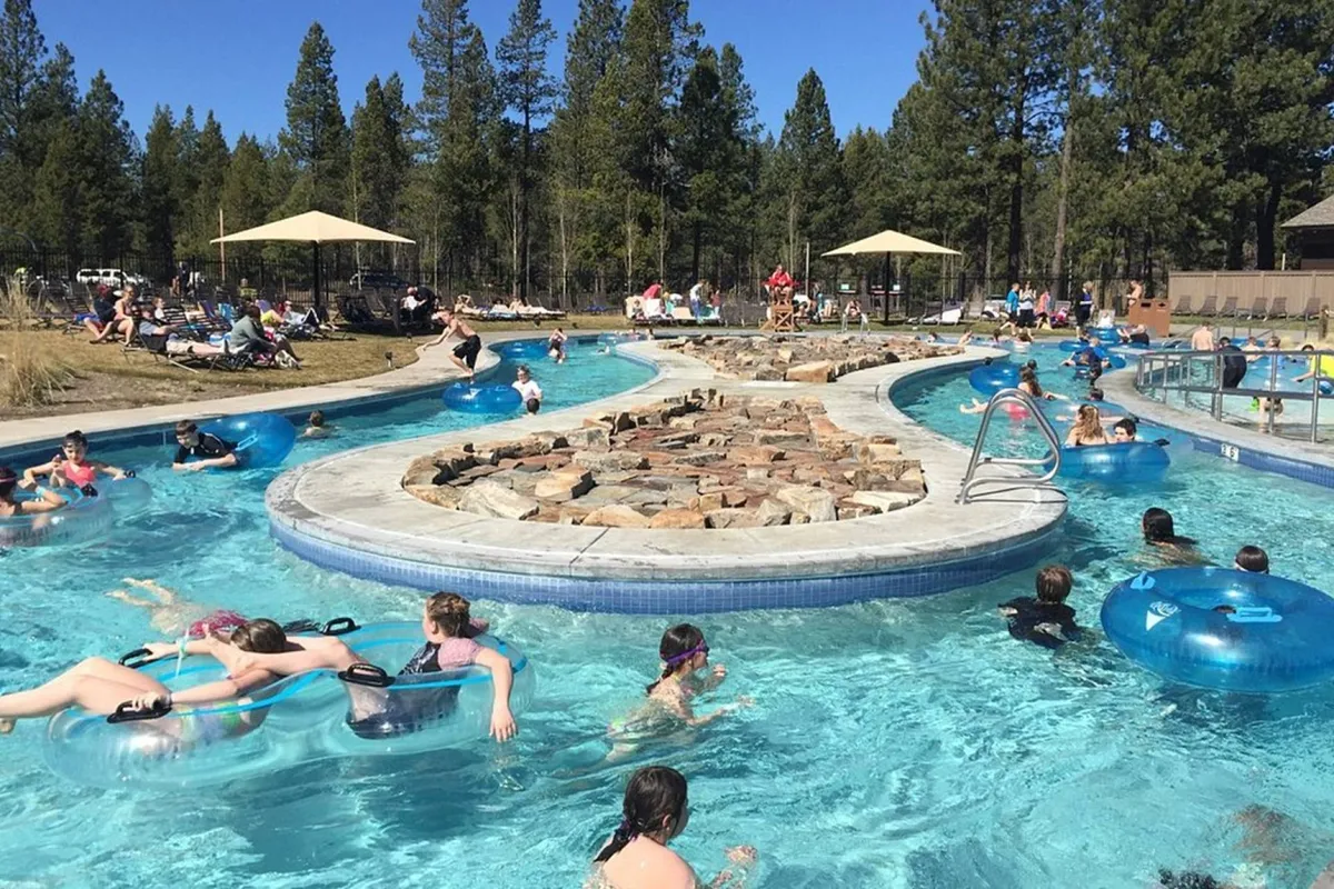 A vibrant outdoor pool scene at the Sunriver Aquatic Center, featuring families and friends floating on colorful inflatables, with natural rock formations and tanning areas surrounding the water. The mood is carefree and joyful, emphasizing water play and social interaction.